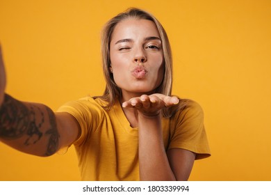 Image of young joyful woman blowing air kiss and taking selfie photo isolated over yellow background