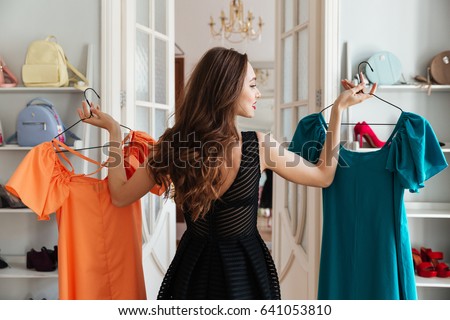 Image of young lady standing in clothes shop indoors choosing dresses. Looking aside.