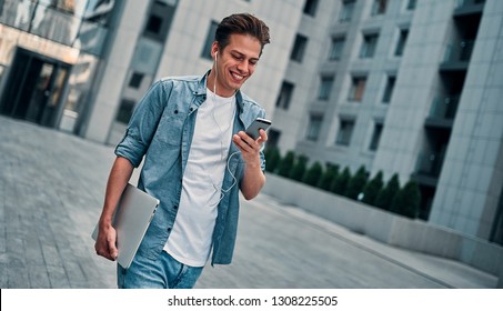 Image of young man, student in headphones walking in the city using mobile phone holding laptop computer. - Shutterstock ID 1308225505