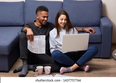 Image of young multicultural couples sitting on floor near blu sofa, using portabl compute for online working, two college students writting course work together, using lap top and wireless Internet.