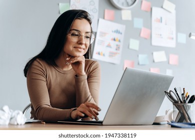 Image Of Young Pleased, Happy, Beautiful Business Woman Sit Indoors In Office Using Laptop Computer. The Girl Wears Glasses.