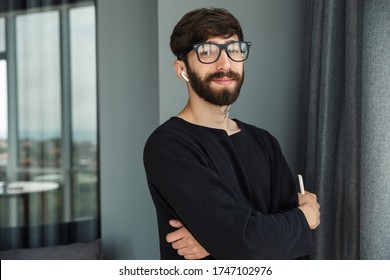 Image of young pleased man holding cellphone and using wireless earphones while posing in living room - Shutterstock ID 1747102976