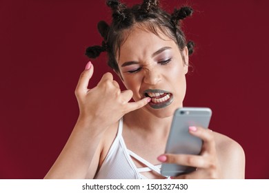 Image Of Young Punk Girl With Bizarre Hairstyle And Makeup Cleaning Her Teeth While Looking At Smartphone Isolated Over Red Background