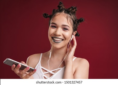 Image Of Young Punk Girl With Bizarre Hairstyle And Dark Lipstick Laughing While Using Smartphone With Earphones Isolated Over Red Background