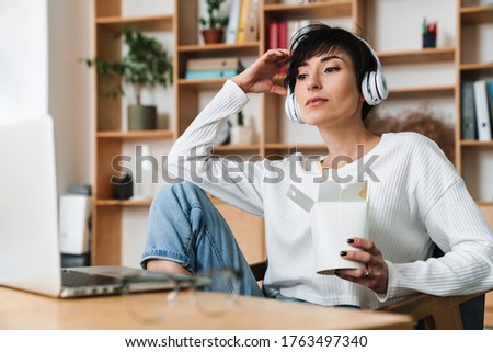 Image of young serious woman in headphones and eating asian noodles while working with laptop indoors