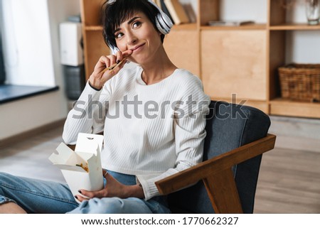Image of young smiling woman using headphones while eating asian noodles takeaway indoors