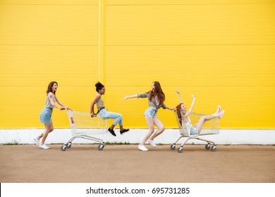 Image of young smiling women friends over yellow wall. Have fun with shopping trolley.
