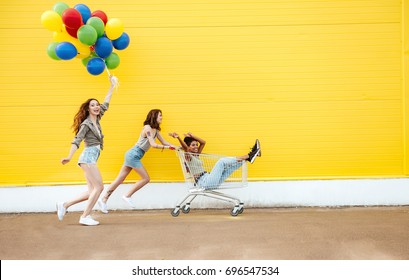 Image of young smiling women friends over yellow wall. Have fun with shopping trolley and balloons. - Powered by Shutterstock