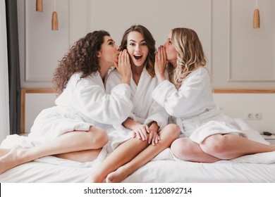 Image of young three women 20s wearing bathrobe sitting on big bed and telling gossips to each others during bridal shower in cozy apartment or fashionable hotel room