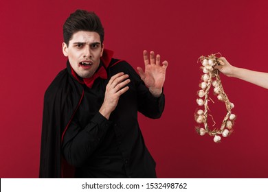 Image Of Young Vampire Man With Fangs In Black Halloween Costume Being Scared Of Garlic Isolated Over Red Wall