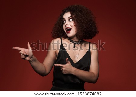Image of young witch girl in black dress with halloween makeup pointing finger aside at copyspace isolated over red wall