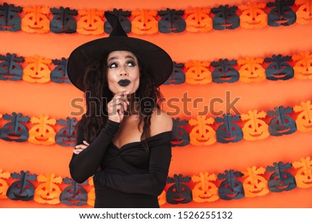 Image of young witch woman in black halloween costume thinking and looking upward isolated over orange pumpkin wall