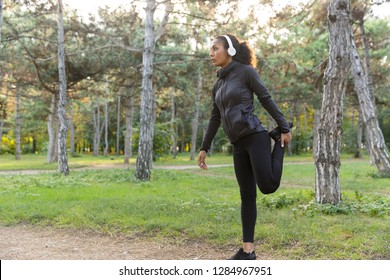 Image of young woman 20s wearing black tracksuit doing sports and stretching body in green park - Powered by Shutterstock