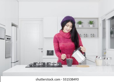 Image Of Young Woman Is Wearing Winter Clothes While Pouring Coffee Into A Mug In The Kitchen