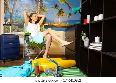 imitation of beach vacations in quarantine. Interiors, room. Young woman on a background of a poster with a sea beach and palm trees. Beach accessories and a suitcase nearby
