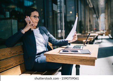 Immersed Calm Interested Lady Looking At Business Paper Attentively During Phone Conversation While Working Remotely With Devices At Cafe Table Outside