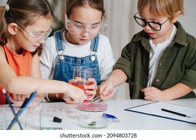 Immersed Little Kids Doing Home Science Project, Filling Glass Dish. All Behid Table, Wearing Glasses. Chemical Glassware And Colored Liquids On The Table.