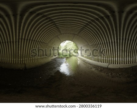 Immersive 360-degree panoramic view from inside a corrugated metal drainage tunnel carrying a shallow stream beneath a roadway. The industrial tunnel structure creates striking geometric patterns and symmetry, while sunlight illuminates the tunnel exit, revealing lush green vegetation outside. Ideal for themes of infrastructure, engineering, transportation, water management, urban development, and environmental design.