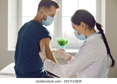 Immunization campaign concept. Side view of female nurse or doctor giving Covid-19 vaccine to male patient. Young man in medical face mask getting flu shot in sunny office of modern clinic or hospital