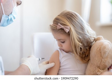 Immunization For Children Concept. Little Cute Blonde Girl Holding A Toy And Getting A Flu Shot Not Afraid Of The Syringe Needle. Doctor Injecting Brave Child With Covid-19 Vaccine At Clinic Or Home