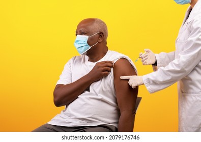 Immunization And Health Care. Senior Black Male Patient In Face Mask Receiving Covid-19 Vaccine Injection On Orange Studio Background. Medical Worker Vaccinating Elderly Man From Coronavirus