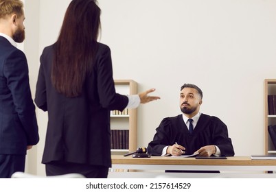 Impartial Judge Listens To Defendant And His Lawyer Who Provides Evidence Of Innocence To Crime. Rear View Of Female Lawyer And Male Defendant Talking To Serious Male Judge Sitting At Table.