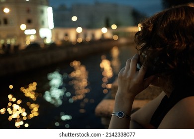 Impersonal Close-up Photo Of A Girl's Head Against The Backdrop Of A River Embankment And City Lights In The Evening Time. She Holding A Mobile Phone To Her Ear With Her Left Hand. Non-personalised 