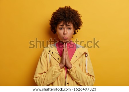 Imploring dark skinned Afro American lady purses lower lips and keeps hands in praying gesture, going to cry as needs help badly, asks for apologise, wears yellow anorak, feels worried and unhappy