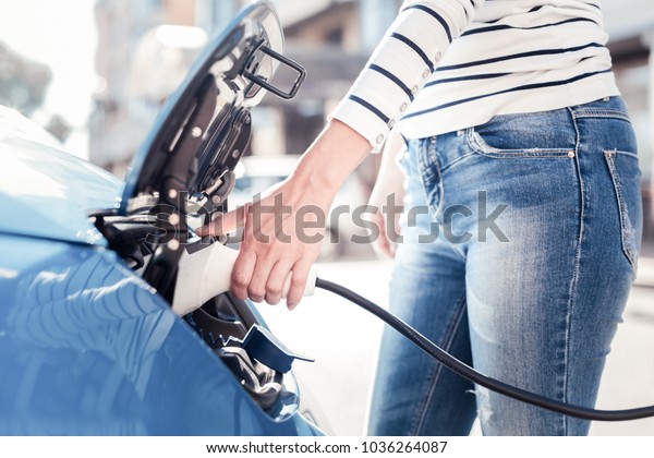 Importance of electricity. Smart young stylish woman
standing on the electric gas station and holding gadget which
charging the car.