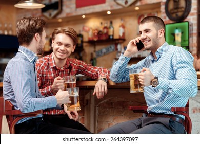 Important call during meeting with friends. Three young men sitting at the bar counter and drinking beer while one of them talks over the phone