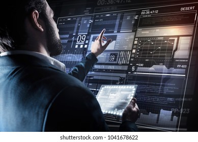 Important changes. Calm interested young programmer thoughtfully pointing to the screen of his futuristic device while holding a tiny tablet and making necessary changes in his new program