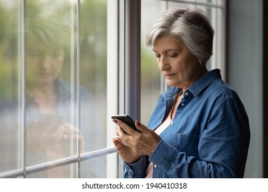 Important Message. Serious Aged Latin Female Pensioner Stand By Picture Window At Home Hold Cell Text In Messenger App. Focused Older Woman Thinking On Email Received From Bank Write Answer. Copyspace