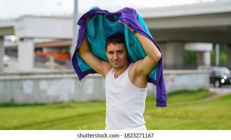 Imposing athletic man in white undershirt putting on jacket on green park lawn