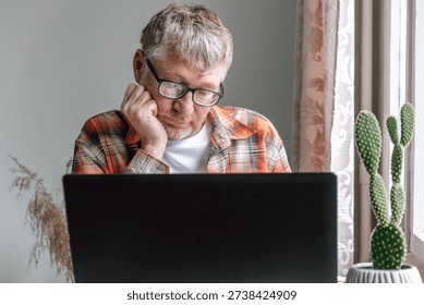 imposing elderly man sits at table, looking at his laptop with thoughtful expression. he is indoors next to window with plants and cactus nearby. close up.