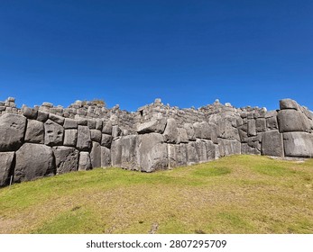 Imposing geometric stone structures form ancient walls against a clear blue sky. The vibrant green of the lawn contrasts with the gray of the rocks.