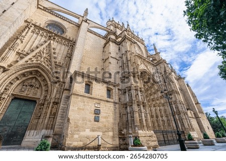 An imposing low-angle view of the intricate Gothic facade of the Seville Cathedral, a UNESCO World Heritage site, showcasing its massive scale and detailed carvings under a partly cloudy sky, Spain.