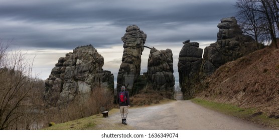 The Imposing Rocks Of The Externsteine Near The Town Of Horn-Bad Meinberg. A Hiker With A Backpack And Walking Sticks Reaches The Monument. There Are Dark Clouds In The Sky.