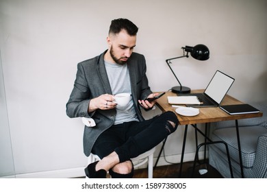 Imposing Self-confident Guy In Ripped Jeans Browsing Phone While Sitting At Desk With Empty Screen Laptop With Raised Leg And Drinking Coffee In Work Room