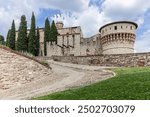 The imposing stone structure of the medieval Brescia Castle in Italy, surrounded by tall cypress trees, captures the historic architecture and serene pathways of this ancient fortification