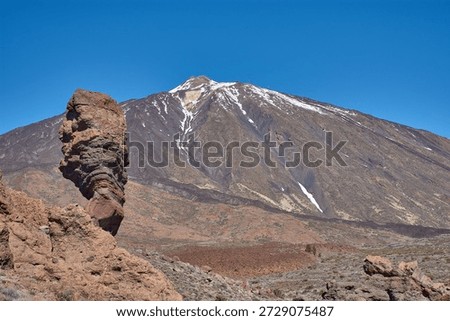 The imposing vertical structure of Roque Cinchado dominates the foreground while the majestic snow-capped peak of Mount Teide rises majestically in the distance. The striking contrast between the ochre hues of the basalt rock, the white of the summit, and the intense blue of the Canary Island sky creates a visually powerful image, ideal for tourism campaigns, geography books, and travel advertising