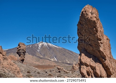 The imposing vertical structure of Roque Cinchado dominates the foreground while the majestic snow-capped peak of Mount Teide rises majestically in the distance. The striking contrast between the ochre hues of the basalt rock, the white of the summit, and the intense blue of the Canary Island sky creates a visually powerful image, ideal for tourism campaigns, geography books, and travel advertising