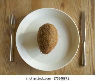 An Impossible Task. A Coconut On A White Dinner Plate With Cutlery On The Sides.