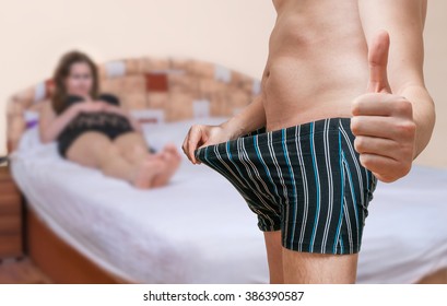 Impotence Concept. Woman Is Waiting In Bed On Her Lover Who Is Looking On His Penis After Viagra And Shows Thumb Up That He Is Ready For Sex.