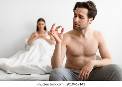 Impotence Issue. Young Husband Holding Potency Pill Before Sex While Wife Waiting In Bed Indoors. Medicine For Male Sexual Health Problems. Low Libido Treatment Concept. Selective Focus