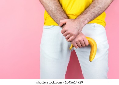 Impotence and men's health. A man in white jeans, legs apart, holds a limp banana near the genitals. Pink background. Close up. Copy space