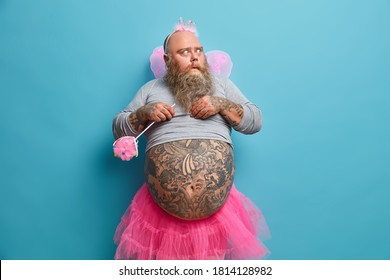 Impressed bearded adult male fairy holds magic wand and stares bugged eyes, wears undersized shirt skirt wings. Man holiday performer looks stupefied aside, isolated on blue studio background