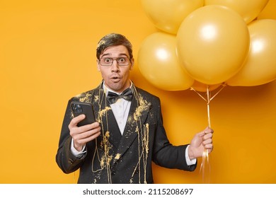 Impressed dark haired man wears festive clothes holds mobile phone and bunch of helium balloons celebrates special occasion has party isolated over yellow background. People holidays concept