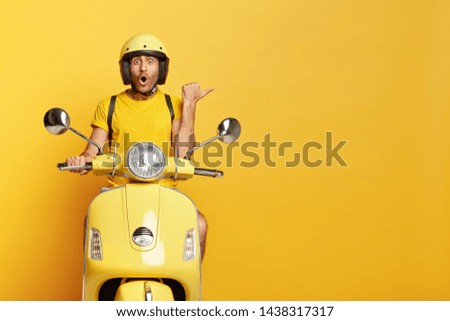 Impressed driver covers distance on yellow motorbike, wears helmet, indicates with great wonder aside, stops on road, demonstrates blank space for your advertising content, tries new vehicle.