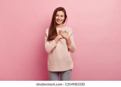 Impressed friendly pleasant looking woman keeps hands on chest, touched by compliment, smiles positively, wears jumper and jeans, models over pink background, looks at camera with great pleasure