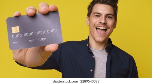 Impressed handsome young person holds in hand debit credit card happy open mouth isolated on yellow color background. Close up of big bank card in hand of 20s young man hipster in blue shirt